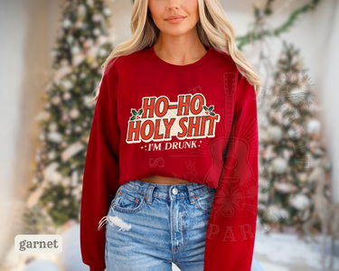 A woman wearing a red sweater with a humorous text printed on it, standing in front of a Christmas tree.
